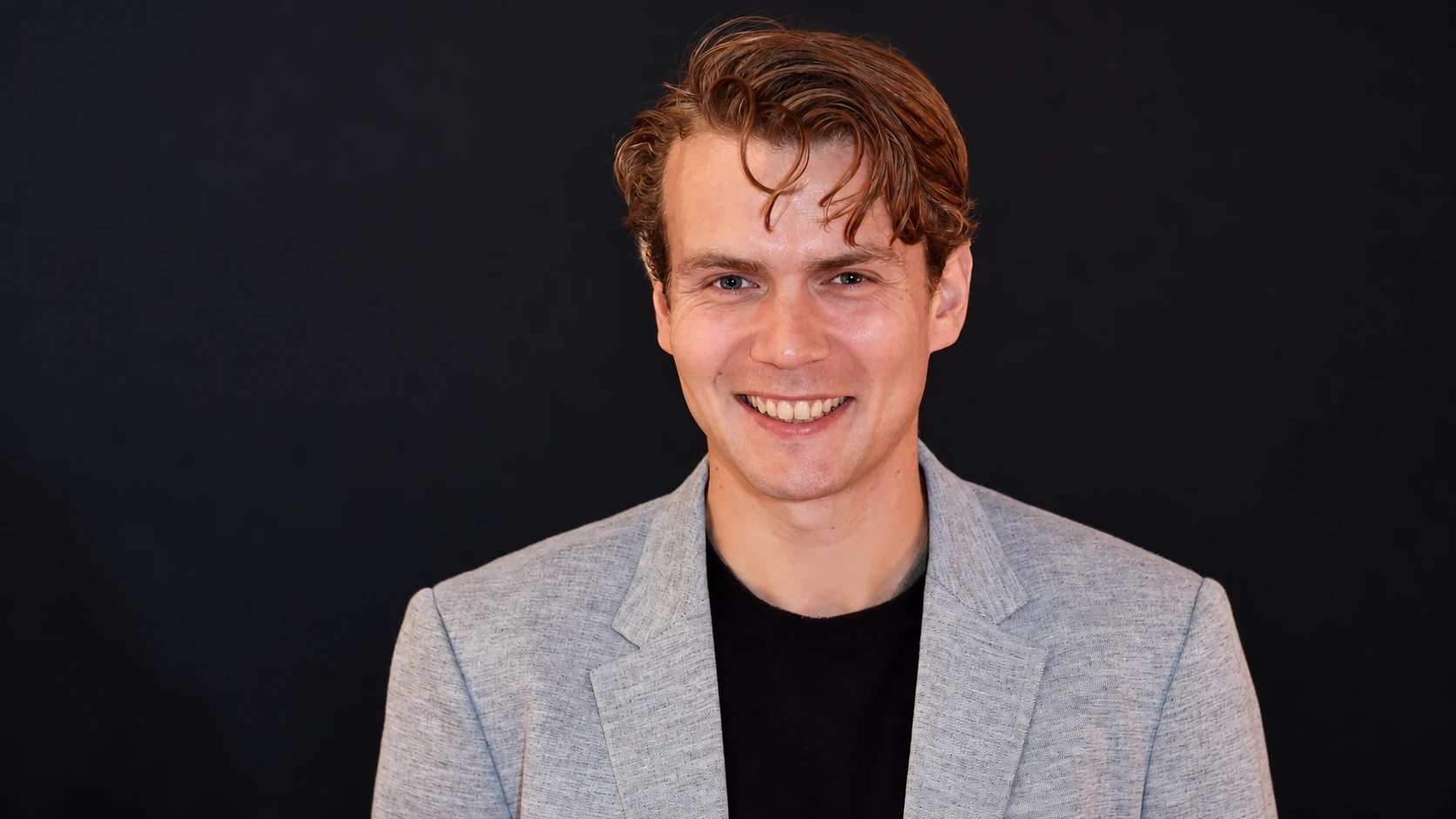 George Farmer smiling in a grey blazer against a dark background