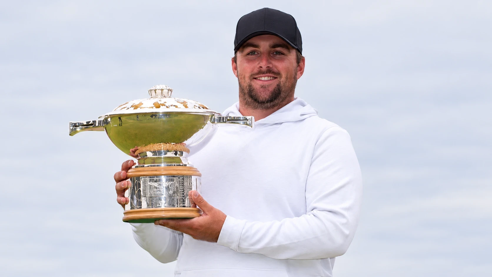 Chris Gotterup in a black cap and white hoodie holding a trophy
