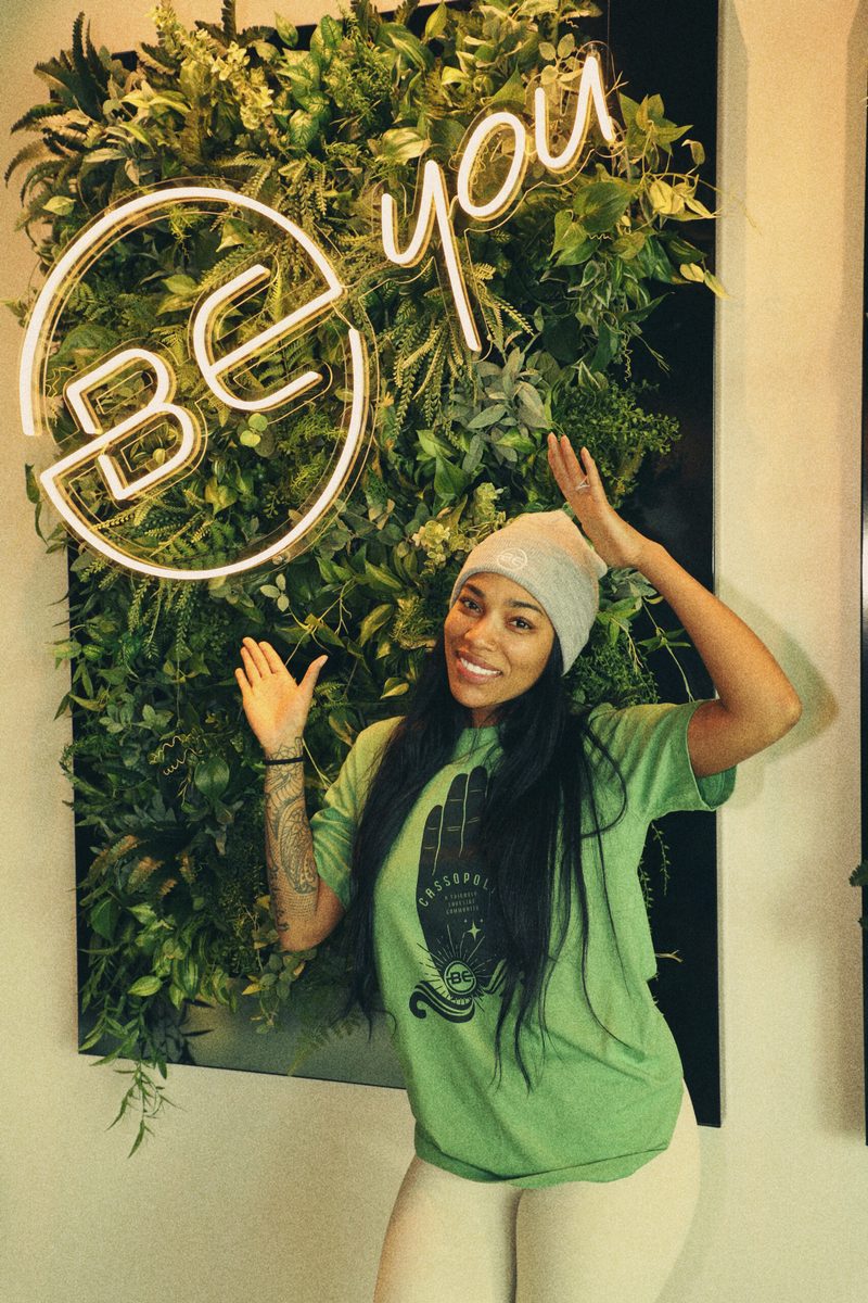 Dreka Gates posing beside a neon 'Be You' sign on a plant wall