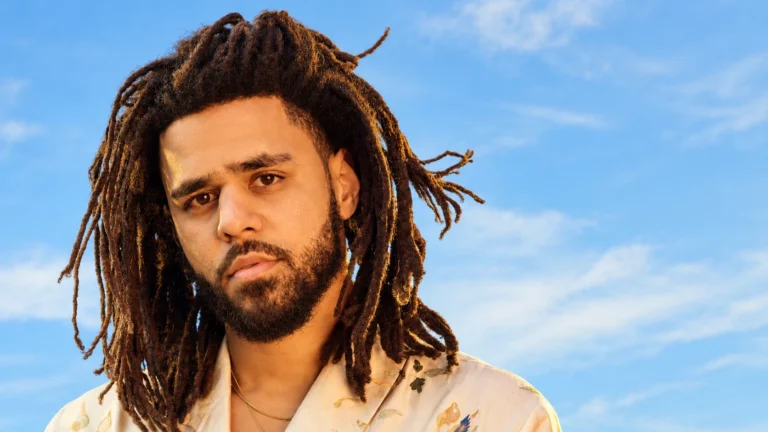 J. Cole with long dreadlocks, posing outdoors under a blue sky